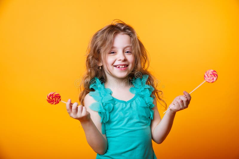 Funny child with candy lollipop, happy little girl eating big sugar lollipop on yellow bright background