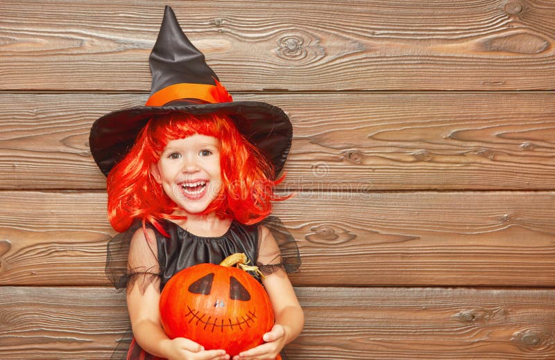 Funny child girl in witch costume for Halloween with pumpkin Jack on a dark wooden background