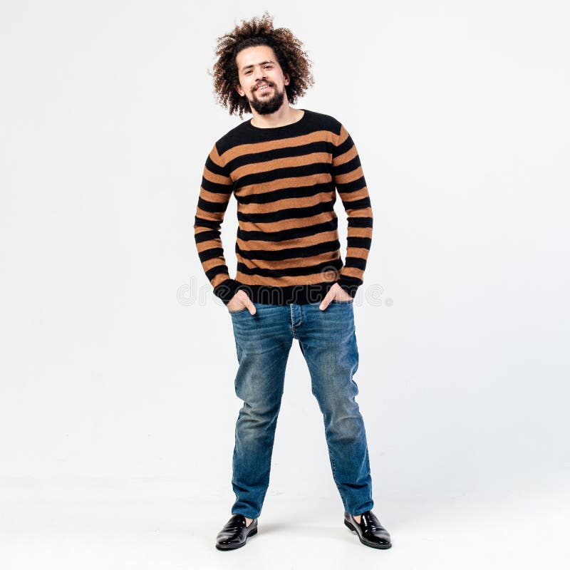 Funny curly guy with a beard dressed in a striped black and yellow sweater and jeans poses in the studio on the white stock image Funny curly guy with a beard dressed in a striped black and yellow sweater and jeans poses in the studio on the white stock image
