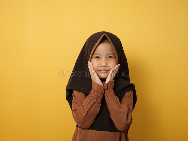 Funny cute Asian muslim little girl wearing hijab looking at camera and smiling, against yellow stock photography