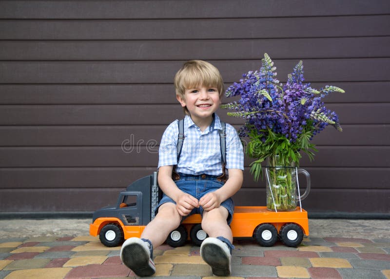 Funny cute 4-5 year old boy in shorts and a shirt sits on a toy truck with a large bouquet of lupins flowers