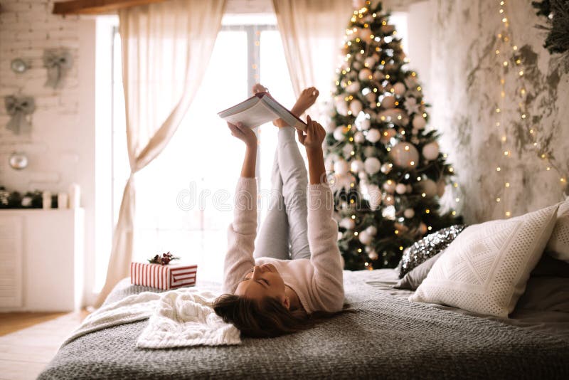 Funny girl dressed in white sweater and pants reads a book liying on the bed with gray blanket, white pillows and a New royalty free stock photo Funny girl dressed in white sweater and pants reads a book liying on the bed with gray blanket, white pillows and a New royalty free stock photo