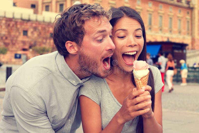 Funny playful young couple eating ice cream. Goofy portrait of boyfriend teasing girlfriend biting off a cold dessert in city summer, Stockholm, Sweden, Europe.