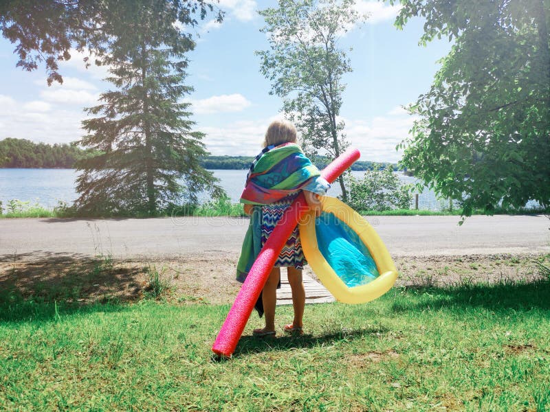 Funny preschool girl with many beach towels and swimming noodle standing near lake river ready to swim on summer day stock photo Funny preschool girl with many beach towels and swimming noodle standing near lake river ready to swim on summer day stock photo