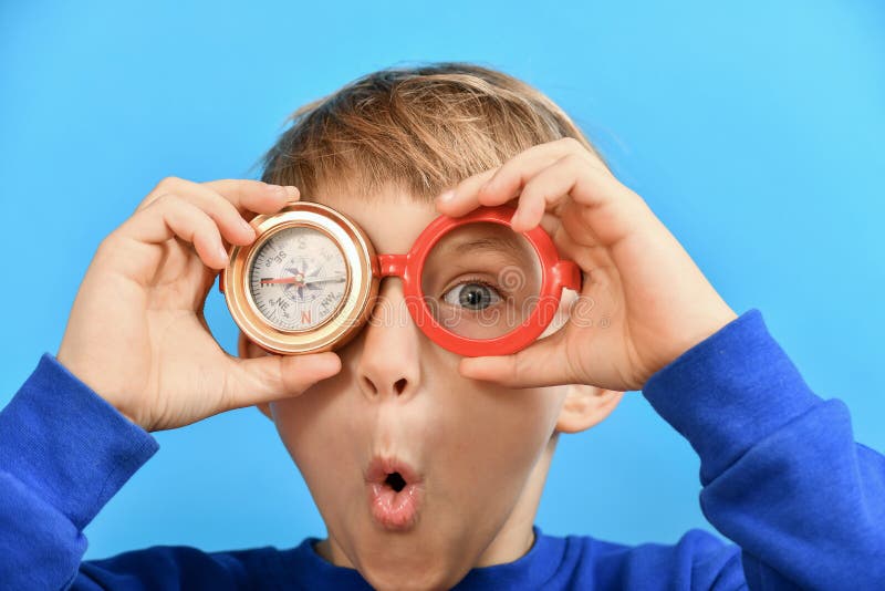 A funny and surprised boy holds a compass in one hand near his eyes and glasses in the other