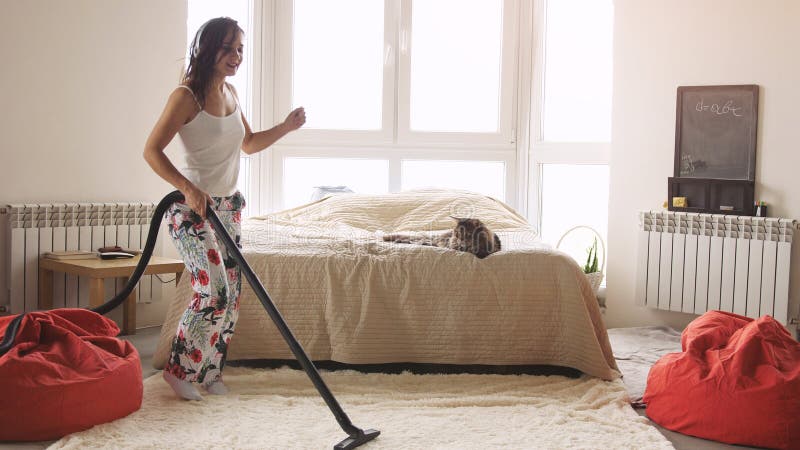 Young woman dancing with vacuum cleaner doing chores cleaning house having fun silly dance listening to music wearing