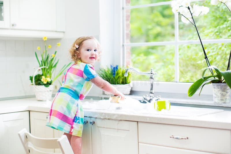 Funny toddler girl in colorful dress washing dishes