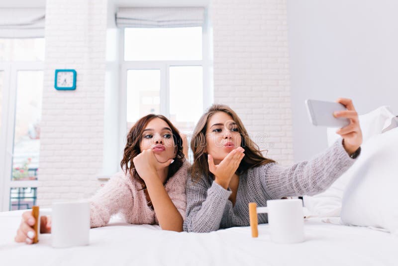 Funny young women in cozy soft sweaters making selfie portrait on bed. Joyful girls having fun, sending a kiss, drinking royalty free stock photography Funny young women in cozy soft sweaters making selfie portrait on bed. Joyful girls having fun, sending a kiss, drinking royalty free stock photography