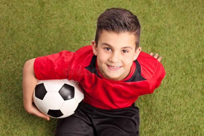 Futbolista menor que se sienta en hierba y la sonrisa fotografía de archivo Futbolista menor que se sienta en hierba y la sonrisa fotografía de archivo