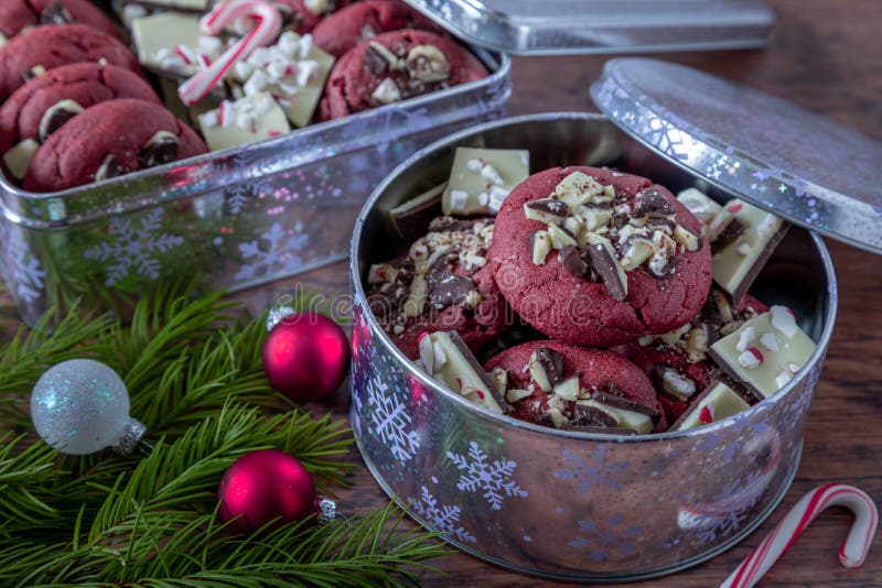 Holiday food gift of peppermint bark stuffed red velvet cookies in a silver cookie tins with snowflakes, small candy canes on wooden table and peppermint bark pieces included with cookies and evergreen branches and ornament decorations. Holiday food gift of peppermint bark stuffed red velvet cookies in a silver cookie tins with snowflakes, small candy canes on wooden table and peppermint bark pieces included with cookies and evergreen branches and ornament decorations