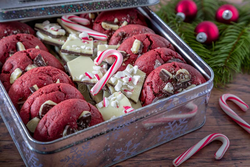 Close up of peppermint bark stuffed red velvet cookies in silver holiday gift tin with peppermint bark candy and candy canes, with evergreen branches and red ornaments. Close up of peppermint bark stuffed red velvet cookies in silver holiday gift tin with peppermint bark candy and candy canes, with evergreen branches and red ornaments