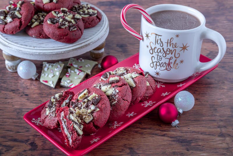 Holiday baking display of peppermint bark stuffed red velvet cookies on a red plate with snowflakes, a cup of hot chocolate with a candy on rim and plate of cookies in background. Holiday baking display of peppermint bark stuffed red velvet cookies on a red plate with snowflakes, a cup of hot chocolate with a candy on rim and plate of cookies in background