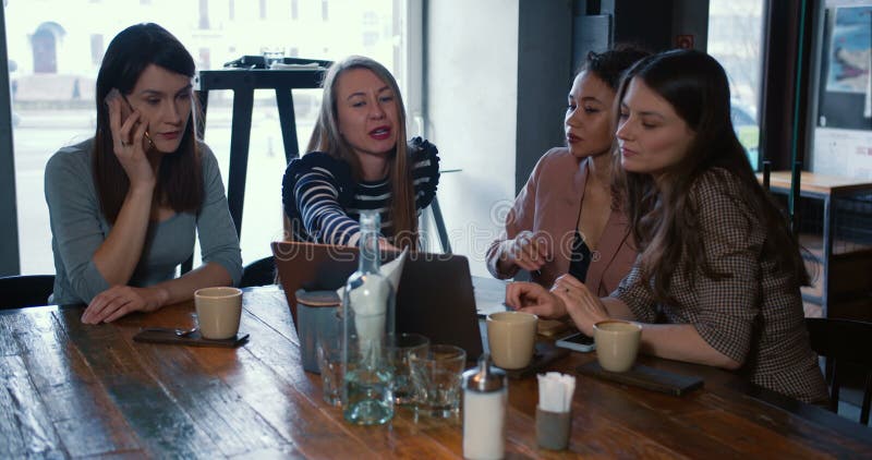 Fyra glada, spännande, vackra, mångskiftande kvinnliga vänner sitter och pratar tillfälligt vid ett modernt kaffepakelmöte. arkivfilmer