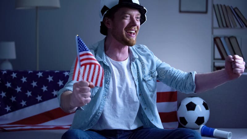 Fã de futebol americano que guarda a bandeira dos EUA, fósforo de futebol de observação na tevê em casa imagens de stock royalty free Fã de futebol americano que guarda a bandeira dos EUA, fósforo de futebol de observação na tevê em casa imagens de stock royalty free