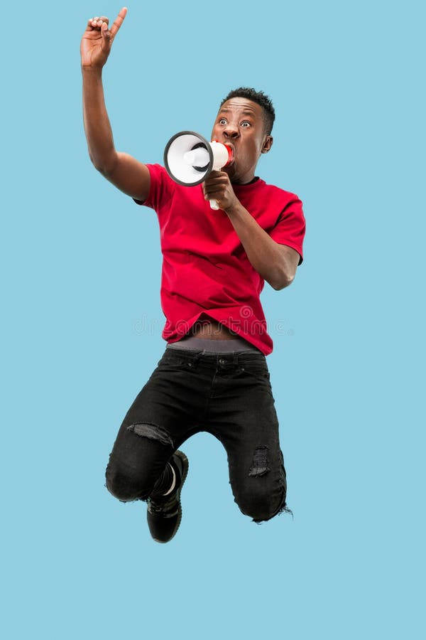 Fã de futebol que salta no fundo azul O homem afro novo como o fan de futebol com megafone fotografia de stock Fã de futebol que salta no fundo azul O homem afro novo como o fan de futebol com megafone fotografia de stock
