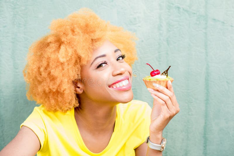 Portrait of a pretty african woman in yellow t-shirt holding a sweet dessert with cherry on the green background. Portrait of a pretty african woman in yellow t-shirt holding a sweet dessert with cherry on the green background