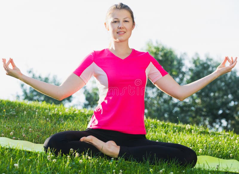 Female 20-30 years old in pink T-shirt is sitting and doing meditation in the park. Female 20-30 years old in pink T-shirt is sitting and doing meditation in the park.