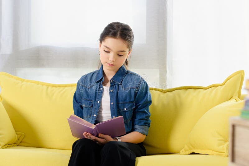 förtjusande preteen barn som sitter på den gula soffan och läseboken arkivfoto förtjusande preteen barn som sitter på den gula soffan och läseboken arkivfoto