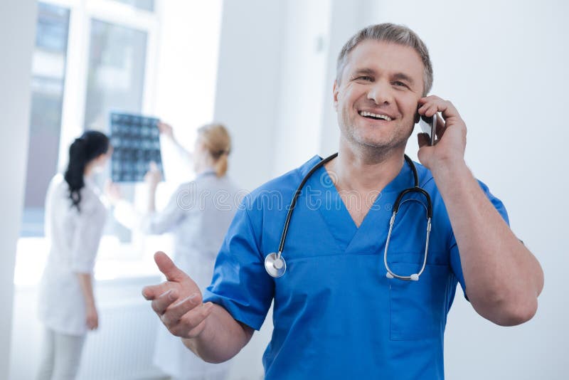 Sharing awesome mood. Charming positive handsome radiologic technologist working at the medical lab and enjoying conversation on the phone while other doctors examining x ray photo in the background. Sharing awesome mood. Charming positive handsome radiologic technologist working at the medical lab and enjoying conversation on the phone while other doctors examining x ray photo in the background