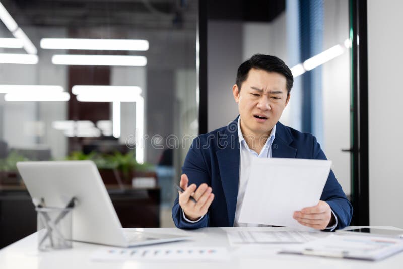 A businessman looks perplexed while examining documents at his work desk in a modern office with bright, linear lighting. A businessman looks perplexed while examining documents at his work desk in a modern office with bright, linear lighting.