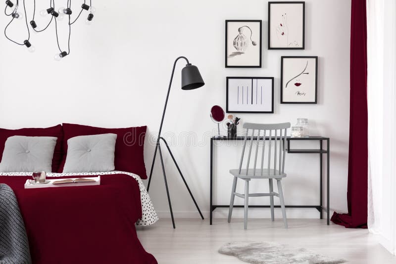Galerij van illustraties op een witte muur boven een klein bureau dat naast een zwarte metaallamp en een bed van Bourgondië in mo stock afbeelding Galerij van illustraties op een witte muur boven een klein bureau dat naast een zwarte metaallamp en een bed van Bourgondië in mo stock afbeelding