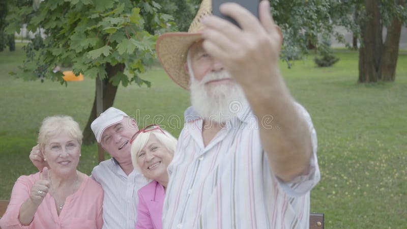 Gamala mannen i en hatt och ett grått skägg gör en selfie mot bakgrunden av hans vänner som sitter på en bänk i, parkerar Tv?