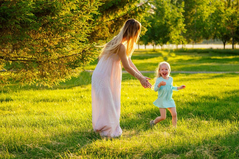 Game of tag and hide and seek .cute little blonde girl in a blue dress with beautiful long-haired mother having fun and playing catch-up in summer day in field greens grass . happy children`s Day.