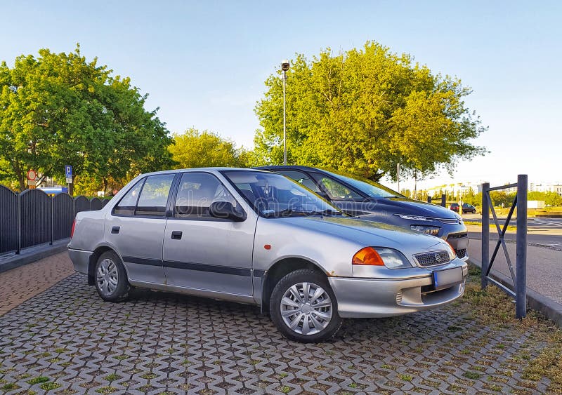 Old popular Japanese small supermini sedan car light silver grey Suzuki Swift 1.3 parked. Second generation introduced in 1988 to 2003. Four door version, right side view and front view with logo. Parked on a street with green trees in the background. Sunny day in May 2022. Old popular Japanese small supermini sedan car light silver grey Suzuki Swift 1.3 parked. Second generation introduced in 1988 to 2003. Four door version, right side view and front view with logo. Parked on a street with green trees in the background. Sunny day in May 2022.