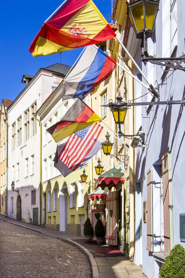 Gammal stenlagd gata med gataljus och flaggor av Tyskland, USA, Ryssland och Spanien som hänger på färgrika hus, Tallinn, Estland arkivbild Gammal stenlagd gata med gataljus och flaggor av Tyskland, USA, Ryssland och Spanien som hänger på färgrika hus, Tallinn, Estland arkivbild