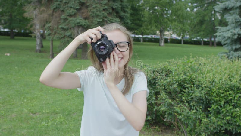 Ganska blonda unga kvinnliga fotografer i glasögon som tar bilder i parken på en varm soldag. Hennes kamera pekade på