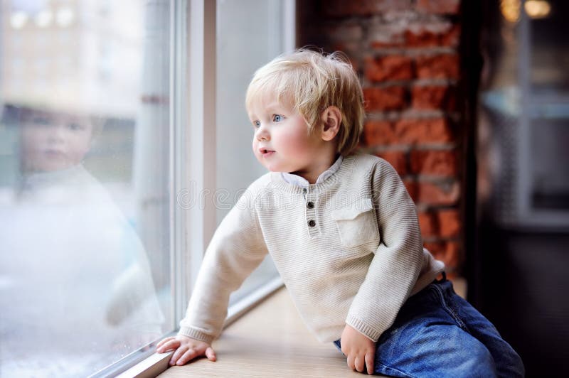 Garçon d'enfant en bas âge s'asseyant sur le filon-couche de fenêtre et regardant la fenêtre photos libres de droits Garçon d'enfant en bas âge s'asseyant sur le filon-couche de fenêtre et regardant la fenêtre photos libres de droits