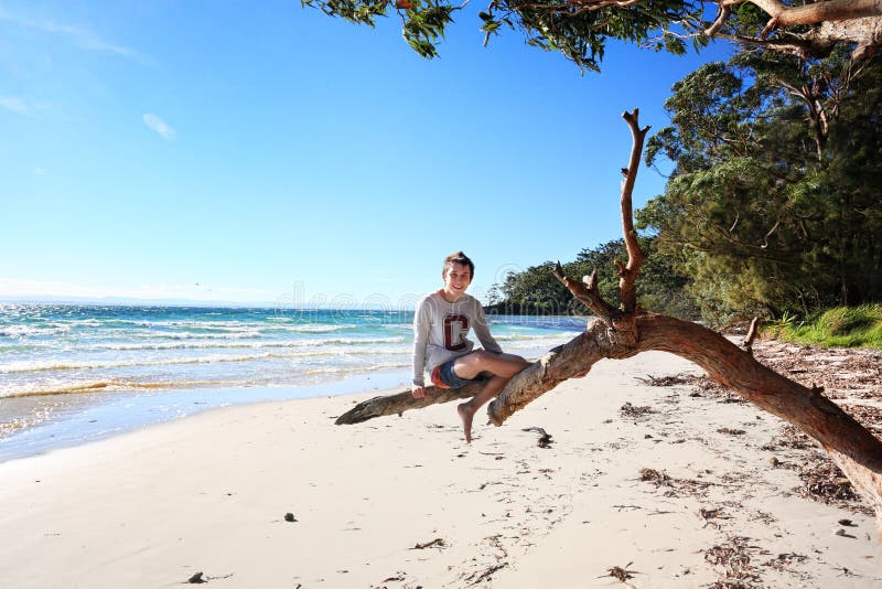 Garçon de l'adolescence gai s'asseyant des vacances d'arbre à la plage Australi