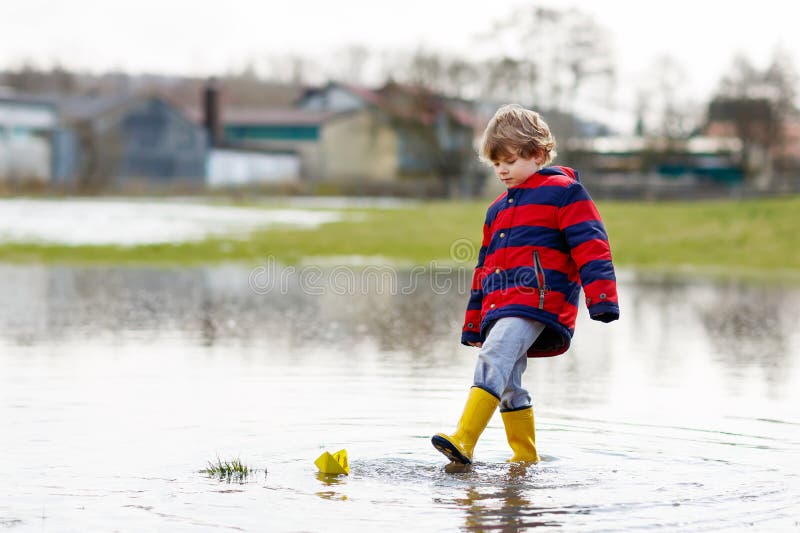 Garçon heureux de petit enfant dans des bottes de pluie jaunes jouant avec le bateau de papier de bateau par le magma énorme le j photo libre de droits Garçon heureux de petit enfant dans des bottes de pluie jaunes jouant avec le bateau de papier de bateau par le magma énorme le j photo libre de droits
