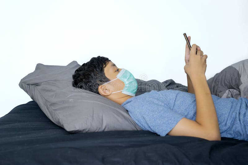 Garçon mignon en utilisant le téléphone portable sur le lit avec le masque image libre de droits