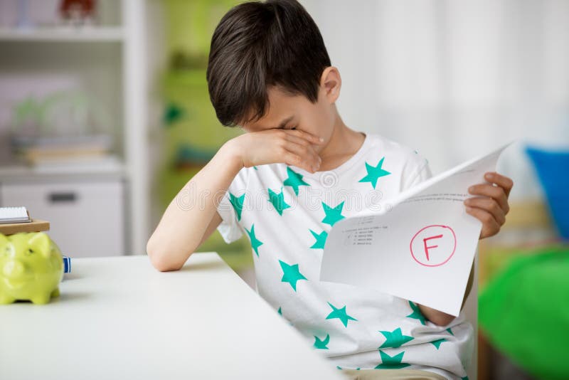 Childhood, education and people concept - sad boy holding school test with f grade. Childhood, education and people concept - sad boy holding school test with f grade