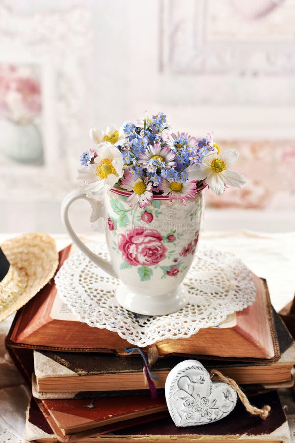 A small bunch of daisy and forget-me-not flowers in a ceramic cup standing on a stack of opened old books in vintage style interior. A small bunch of daisy and forget-me-not flowers in a ceramic cup standing on a stack of opened old books in vintage style interior
