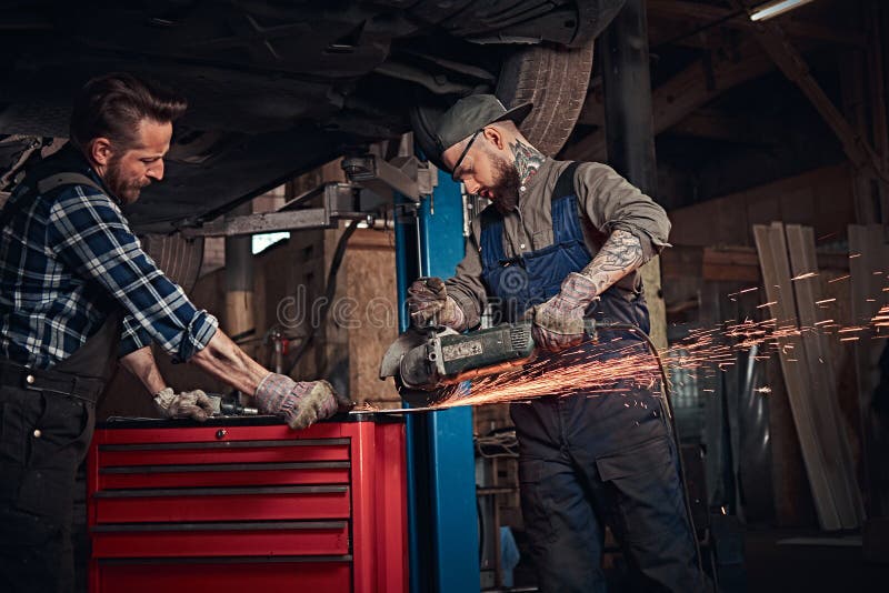 Gebaarde autowerktuigkundige twee in eenvormige en een veiligheidsbril die met een hoekmolen werken terwijl status onder opheffen stock fotografie Gebaarde autowerktuigkundige twee in eenvormige en een veiligheidsbril die met een hoekmolen werken terwijl status onder opheffen stock fotografie