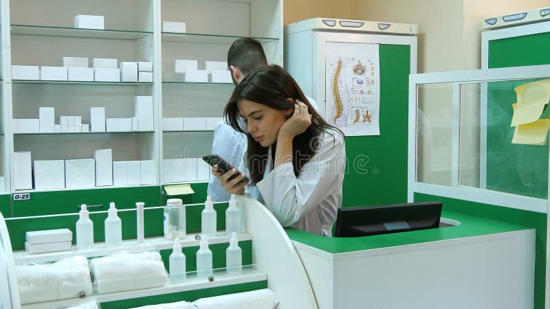 Gebohrter junger Apotheker, der über Handy am Arbeitsplatz broqsing ist lizenzfreie stockbilder Gebohrter junger Apotheker, der über Handy am Arbeitsplatz broqsing ist lizenzfreie stockbilder