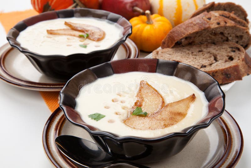 Two bowls of hot delicious Roasted Parsnip and Pear Soup. Garnished with slices of caramelized pears, maple syrup, and parsley. Pumpkins and bread. Two bowls of hot delicious Roasted Parsnip and Pear Soup. Garnished with slices of caramelized pears, maple syrup, and parsley. Pumpkins and bread.