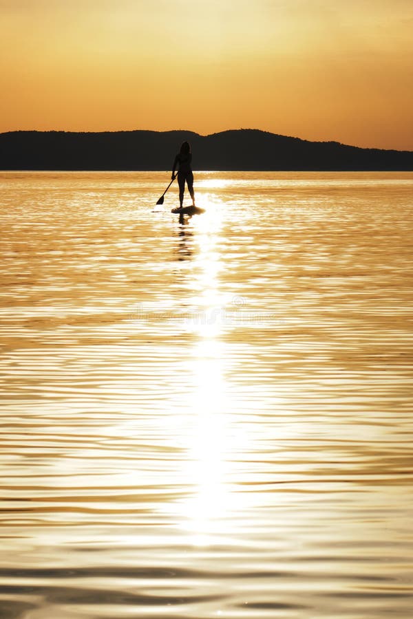 Gecentreerde zonbezinning in water en silhouet van het jonge vrouw paddelen bij zonsondergang op tribune omhoog paddleboard SUP i royalty-vrije stock foto Gecentreerde zonbezinning in water en silhouet van het jonge vrouw paddelen bij zonsondergang op tribune omhoog paddleboard SUP i royalty-vrije stock foto