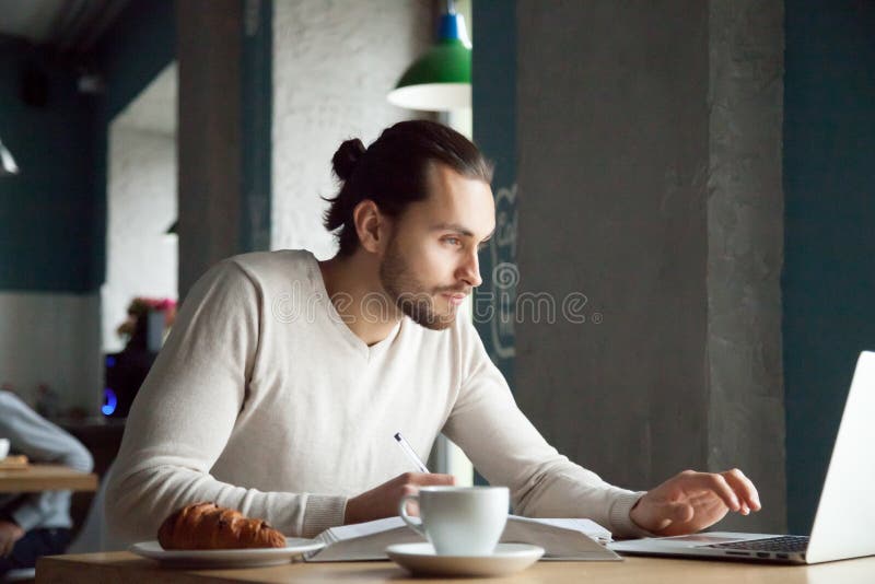 Focused man writing making notes studying with laptop in cafe, serious millennial student, businessman or entrepreneur using computer for e-learning and online education in public place concept. Focused man writing making notes studying with laptop in cafe, serious millennial student, businessman or entrepreneur using computer for e-learning and online education in public place concept