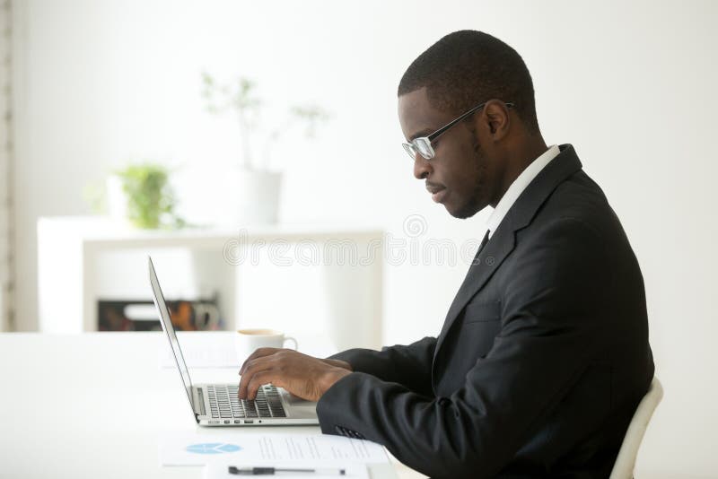 Concentrated self-employed African American manager in glasses and formal wear working at laptop, typing company financial report or reading business email at workplace. Concept of focus, hardworking. Concentrated self-employed African American manager in glasses and formal wear working at laptop, typing company financial report or reading business email at workplace. Concept of focus, hardworking