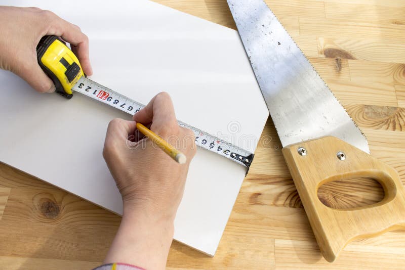 Geerntete Hände einer markierenden weißen Holzverkleidung des Tischlers mit gelbem Bleistift- und Bandmachthaber, Säge liegt in d lizenzfreie stockfotografie Geerntete Hände einer markierenden weißen Holzverkleidung des Tischlers mit gelbem Bleistift- und Bandmachthaber, Säge liegt in d lizenzfreie stockfotografie