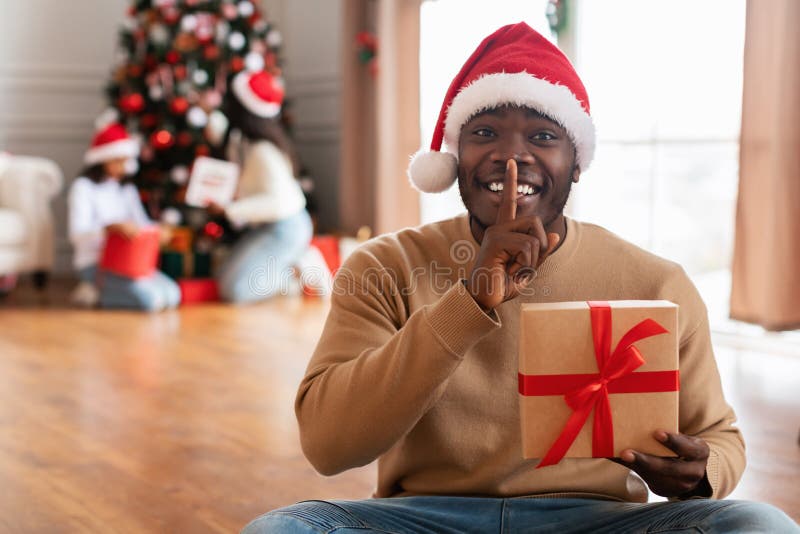 Secret Present. Joyful African American Guy In Santa Claus Hat Holding Gift Box Preparing Surprise For Xmas At Home, Showing Silence Gesture With Finger On Lips Hush Sign, Family Decorating Tree. Secret Present. Joyful African American Guy In Santa Claus Hat Holding Gift Box Preparing Surprise For Xmas At Home, Showing Silence Gesture With Finger On Lips Hush Sign, Family Decorating Tree
