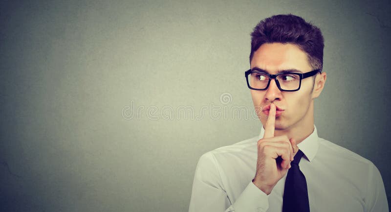 Secret guy. Man saying hush be quiet with finger on lips gesture looking to the side on gray wall background. Secret guy. Man saying hush be quiet with finger on lips gesture looking to the side on gray wall background.