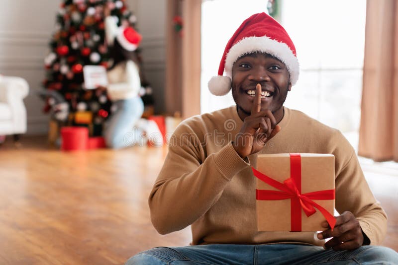 Secret Santa. Portrait Of Excited African American Man In Red Hat Holding Wrapped Box With Present, Making Surprise, Showing Silence Gesture With Finger On Lips While His Girlfriend Decorating Tree. Secret Santa. Portrait Of Excited African American Man In Red Hat Holding Wrapped Box With Present, Making Surprise, Showing Silence Gesture With Finger On Lips While His Girlfriend Decorating Tree