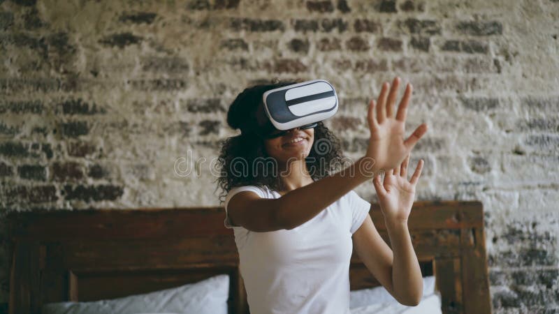 Gelockte Afroamerikanerfrau, die zu Hause Erfahrung unter Verwendung der Kopfhörergläser VR 360 virtueller Realität erhält