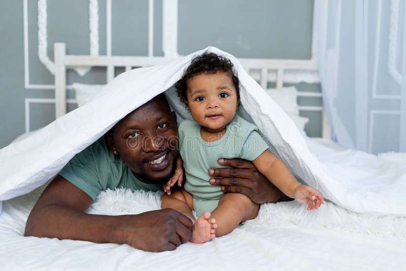Gelukkig glimlachend afrikaanse amerikaanse vader met zoontje op het bed thuis , die onder de deken vrolijke vaders dag zit