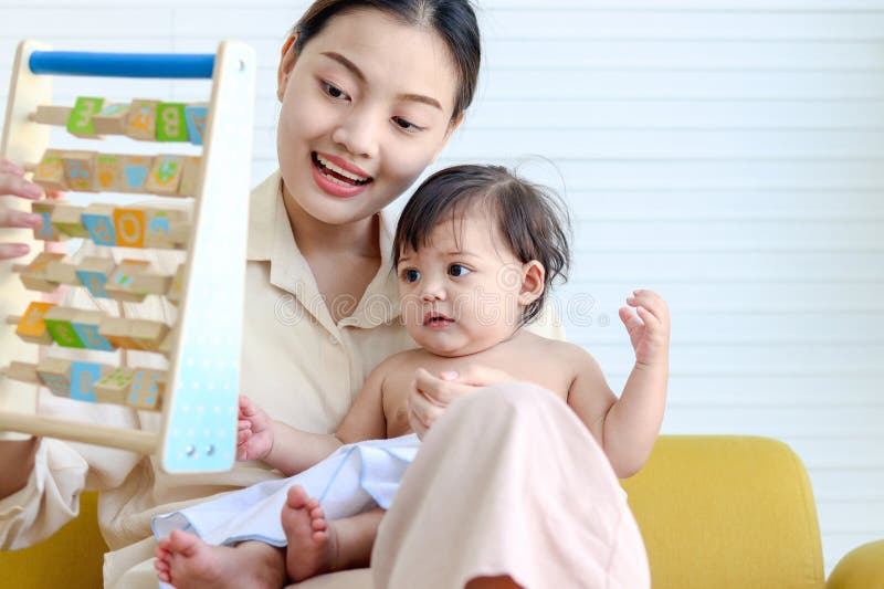 Happy smiling toddle baby sits on mother lap on yellow sofa in living room, little cute kid girl plays with developmental toys, mom plays with daughter child, love in family, childhood and motherhood. Happy smiling toddle baby sits on mother lap on yellow sofa in living room, little cute kid girl plays with developmental toys, mom plays with daughter child, love in family, childhood and motherhood