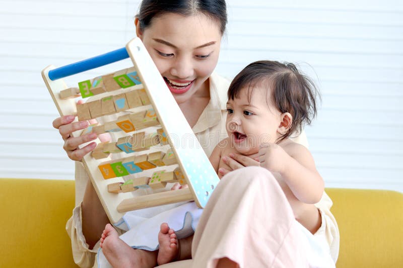 Happy smiling toddle baby sits on mother lap on yellow sofa in living room, little cute kid girl plays with developmental toys, mom plays with daughter child, love in family, childhood and motherhood. Happy smiling toddle baby sits on mother lap on yellow sofa in living room, little cute kid girl plays with developmental toys, mom plays with daughter child, love in family, childhood and motherhood